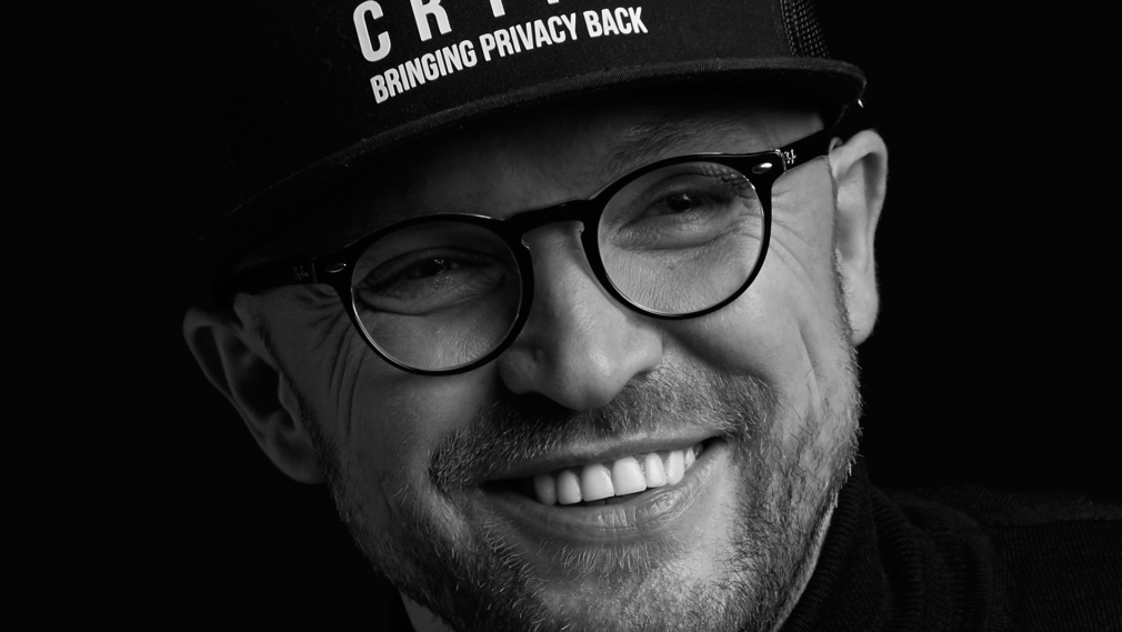 Pawel Makowski pozuje uśmiechnięty w czapce UseCrypt ubrany w okulary czarny golf, zdjęcie jest czarno białe