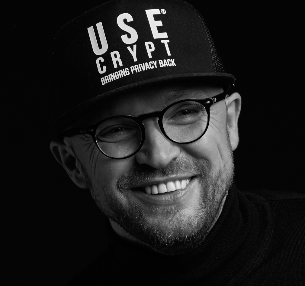 Pawel Makowski pozuje uśmiechnięty w czapce UseCrypt ubrany w okulary czarny golf, zdjęcie jest czarno białe